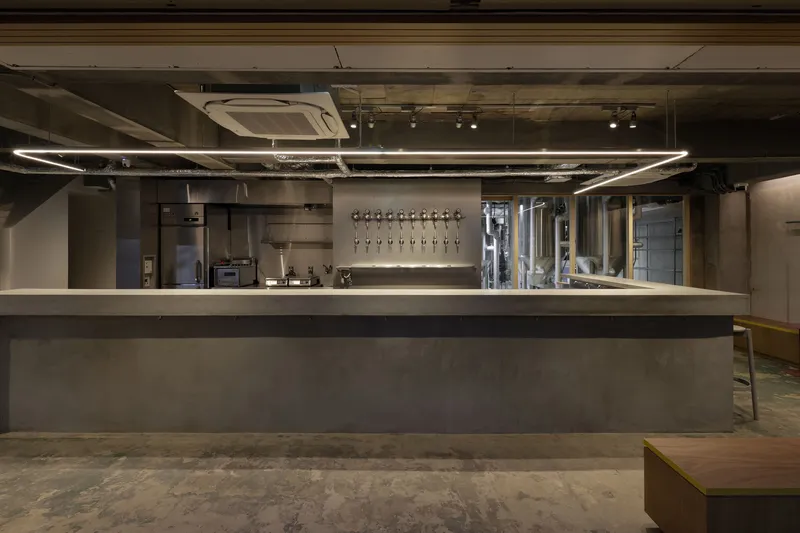 Long concrete bar with tap wall and glass-enclosed fermentation tanks under exposed ductwork