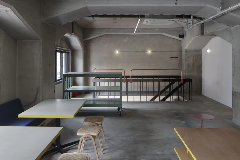 Double-height concrete interior with exposed ductwork, industrial steel railing, and yellow-edged tables