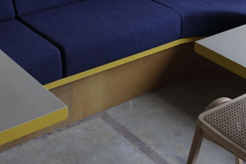 Close-up of yellow metal table edge meeting plywood panel beneath blue upholstered seating