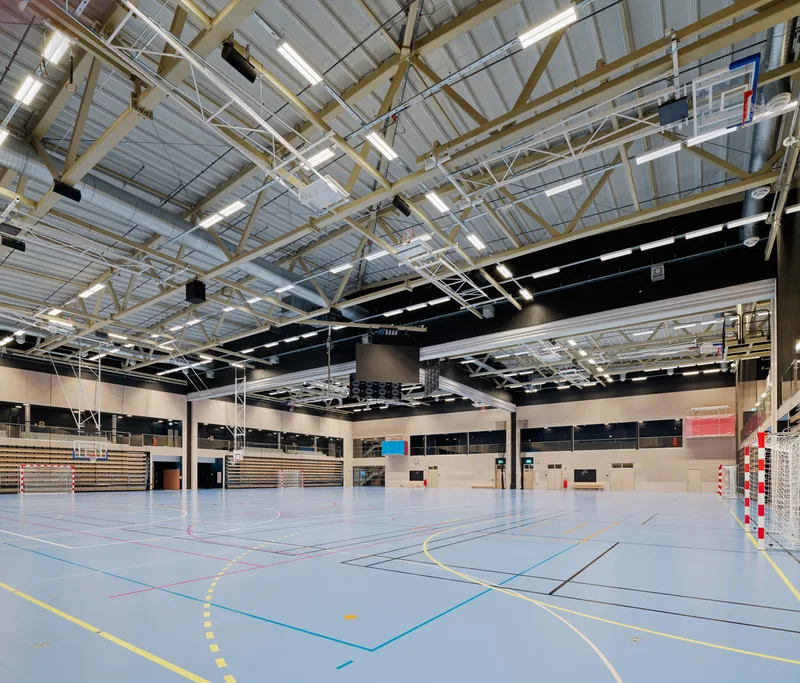 Sports hall with exposed steel trusses and linear tube lights above a multi-line court floor