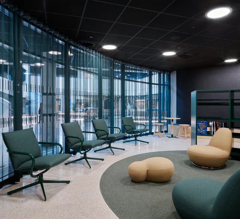 Lounge area with teal armchairs, terrazzo flooring and floor-to-ceiling curtain wall overlooking the campus
