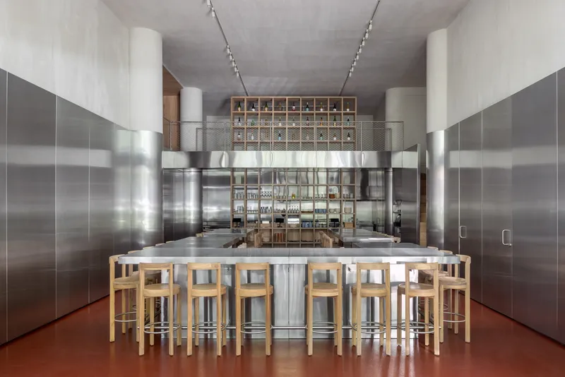 Central island with timber stools surrounded by stainless steel cabinets and a gridded storage wall beyond