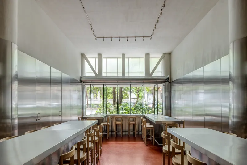 Dining hall with floor-to-ceiling windows overlooking greenery and exposed steel bracing above metal counters