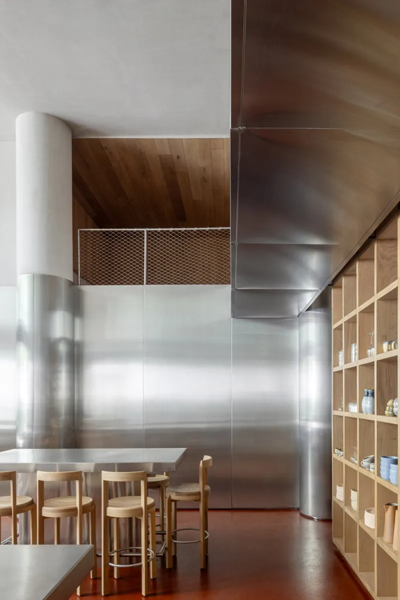 Double-height interior with cylindrical column, timber ceiling and open wood shelving beneath mesh grilles
