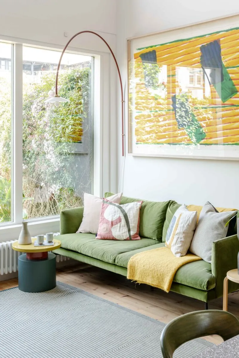 Living room with green upholstered sofa beneath corner windows overlooking garden foliage