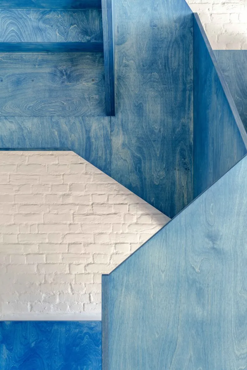 Overlapping planes of blue-stained plywood with white brick wall beneath in shadow