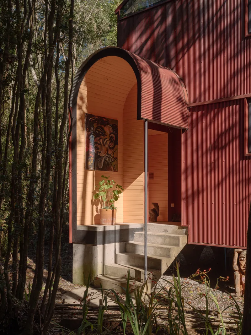 Arched entry portal with concrete stairs and timber handrail beneath corrugated metal canopy in dappled sunlight