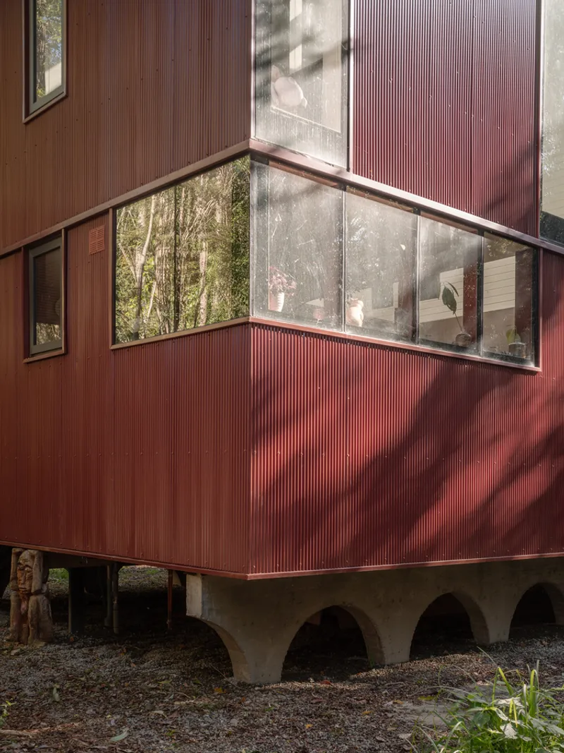 Corner detail of red corrugated metal cladding with glazed bay window above arched concrete foundation