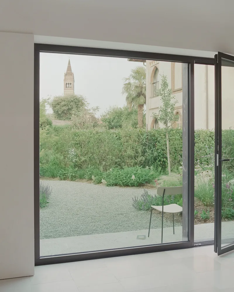 Open sliding glass door framing a view of a church bell tower beyond a garden path