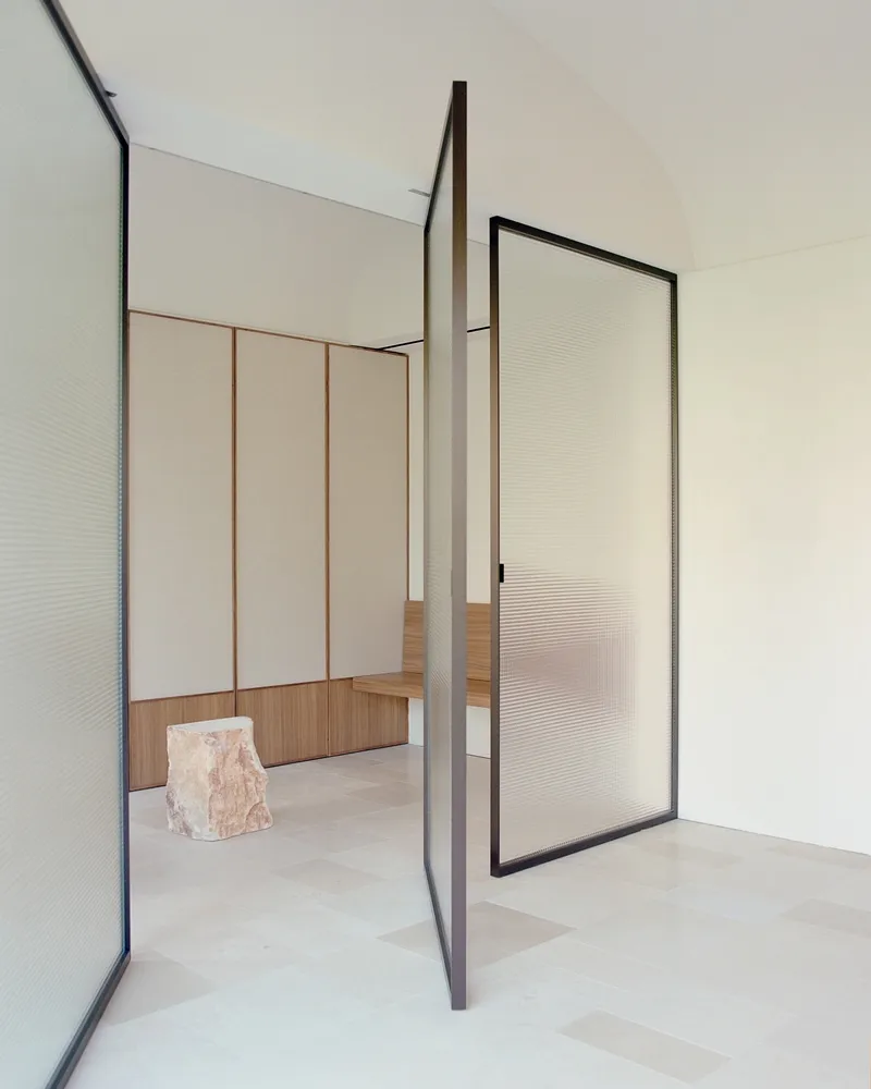 Pivoting translucent glass door framed in dark metal between rooms with pale stone floors