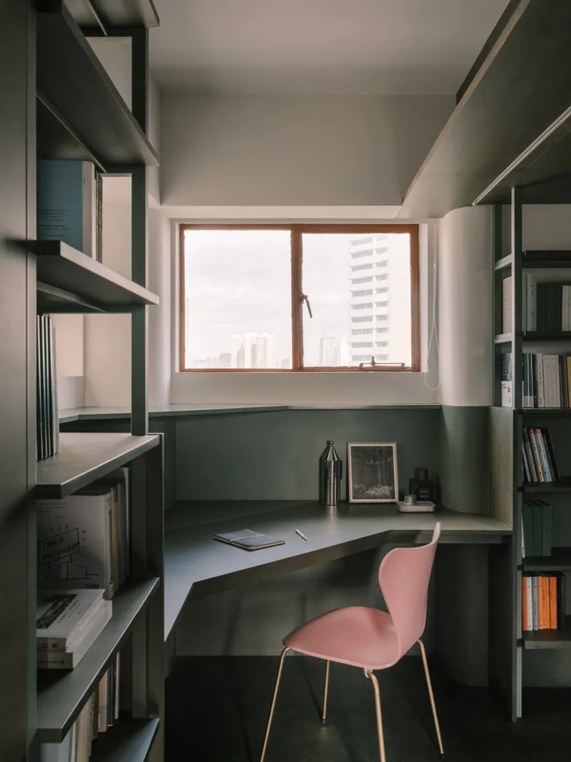 Compact study nook with curved desk, dark green wraparound shelving, and pink chair beneath a timber window