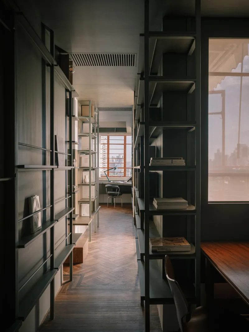 Narrow corridor flanked by open steel shelving units leading to workspace with herringbone parquet flooring