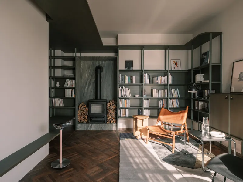 Library with dark green steel shelving, wood-burning stove with firewood storage, and herringbone parquet flooring in afternoon light