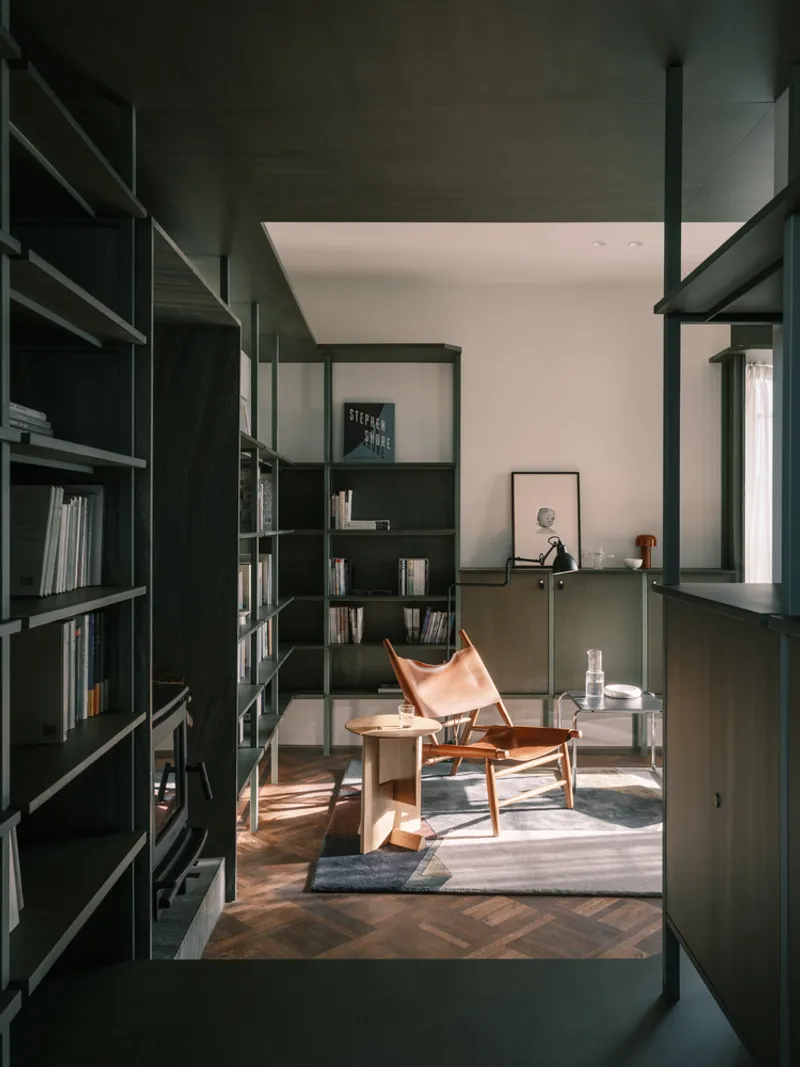 View through dark green steel frame partition into the library with leather sling chair and striped sunlight