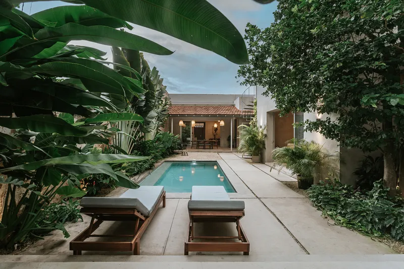 Courtyard pool with concrete deck and wooden loungers framed by banana plants and mature trees at dusk