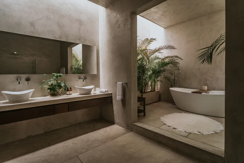 Bathroom with polished concrete surfaces, freestanding tub on a stepped platform, and potted palms beneath a skylight