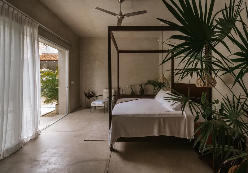 Bedroom with canopy bed frame and polished concrete floors alongside potted palms by sheer curtains