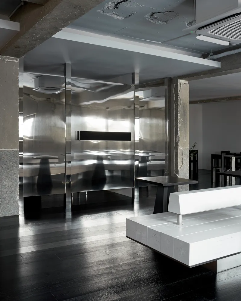 Reflective stainless steel enclosure with transparent glass panels framing a view to the concrete structure beyond