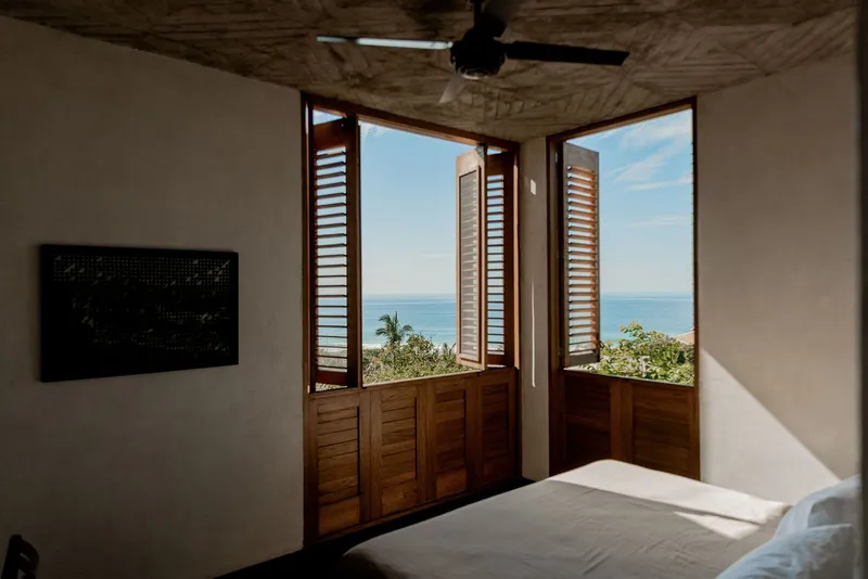 Bedroom with wooden louvered doors opening to an ocean view beneath a woven bamboo ceiling