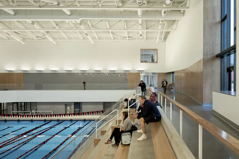 Tiered timber seating along the pool deck with visitors gathering beneath an exposed white ceiling structure