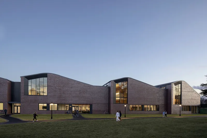 Articulated brick facade with curved roofline and glazed bays illuminated at dusk across a green lawn