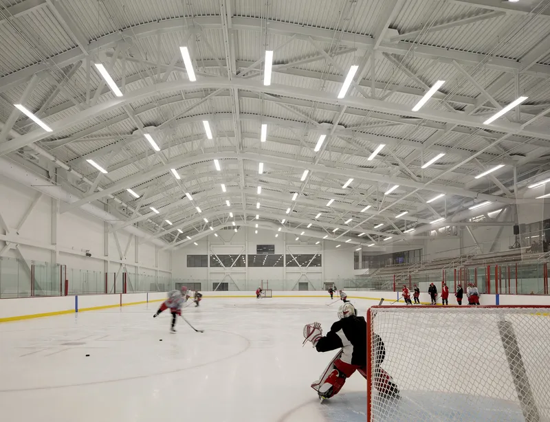 Exposed steel truss ceiling above an indoor ice rink with players and spectators