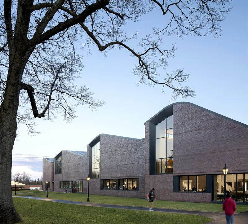 Brick complex with curved rooflines and glazed volumes framed by bare tree branches at dusk