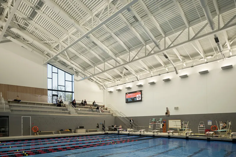 Indoor swimming pool with exposed white steel trusses and tiered seating along one wall