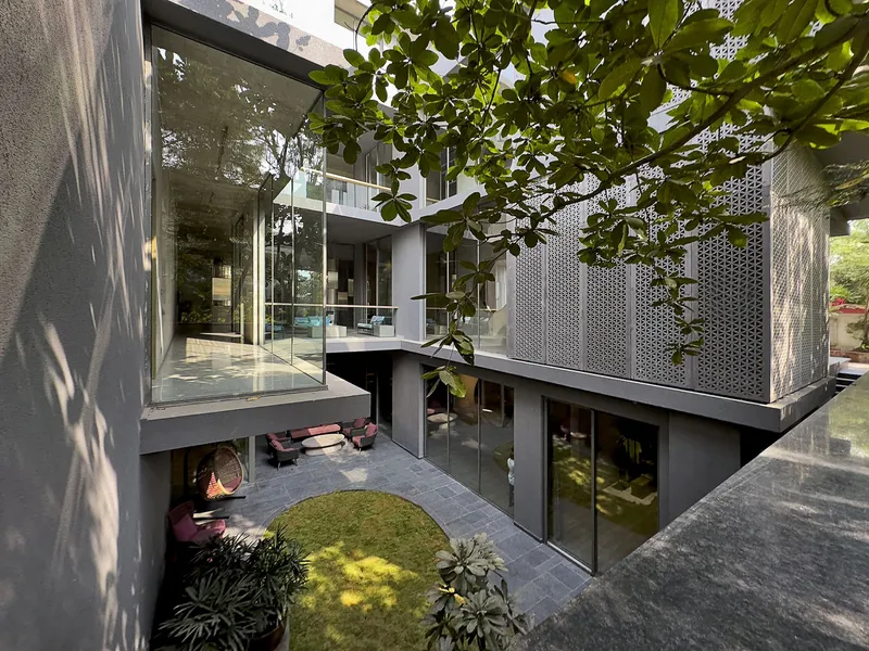 Sunken courtyard with circular grass patch beneath overhanging volumes clad in perforated metal and dappled sunlight filtering through trees