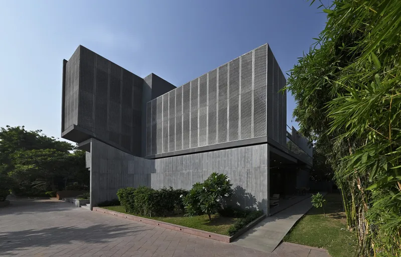 Corner exterior view showing stacked concrete and perforated metal volumes with bamboo and plantings