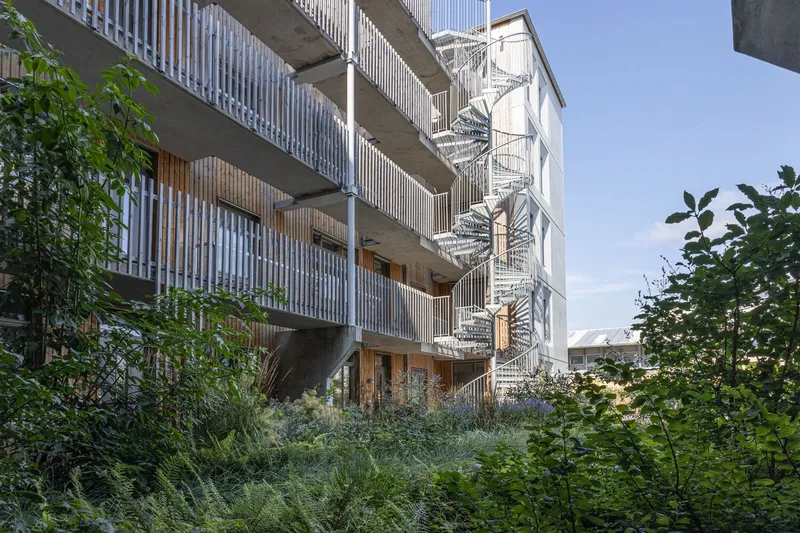 Rear facade showing concrete balconies with vertical timber screening and exterior spiral stair behind planted beds