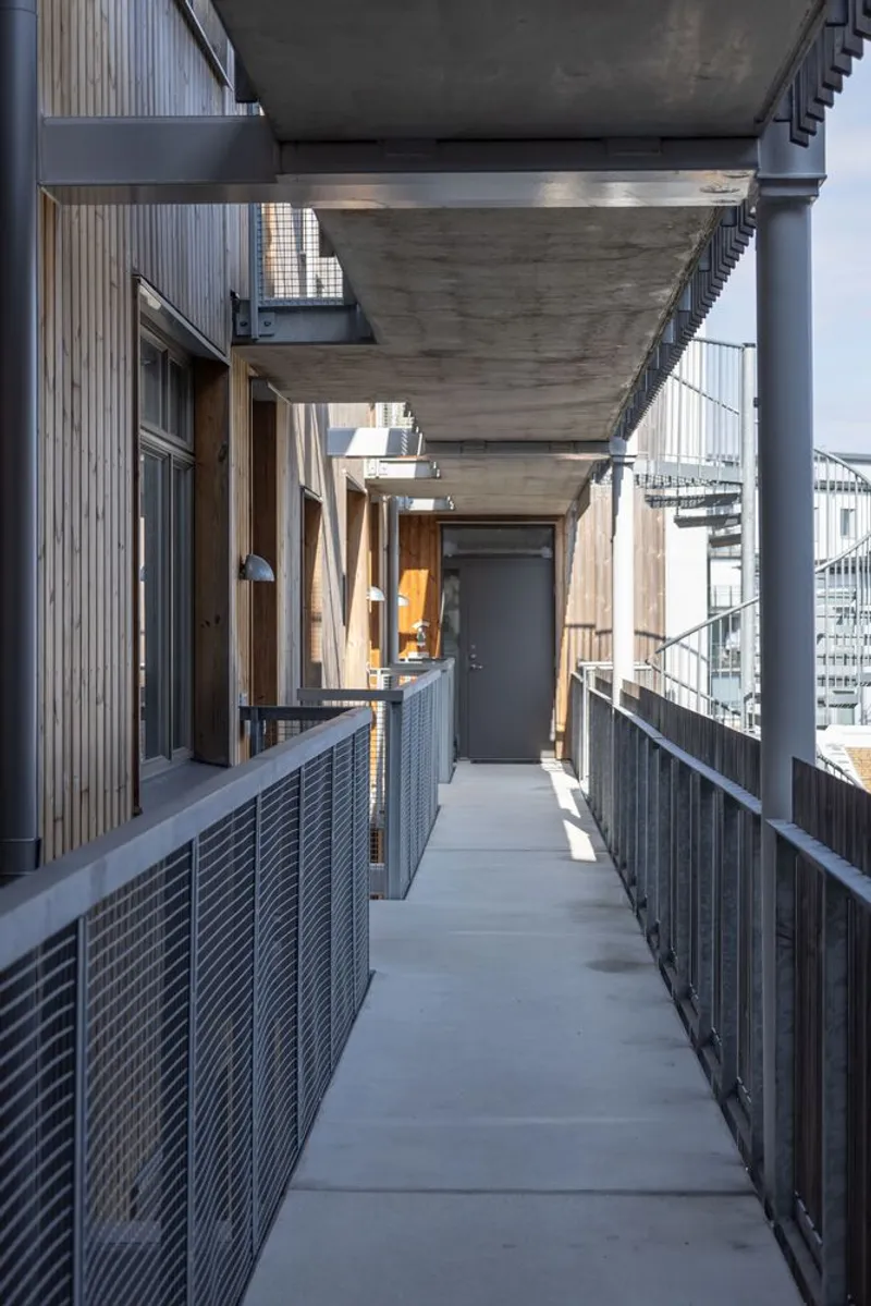 External access corridor with exposed concrete soffits, mesh balustrades, and timber-clad unit entries at dusk