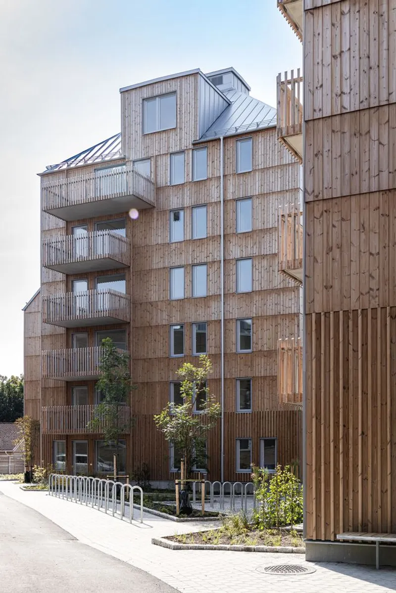 Timber-clad residential block with staggered balconies and metal roofs beside newly planted street trees
