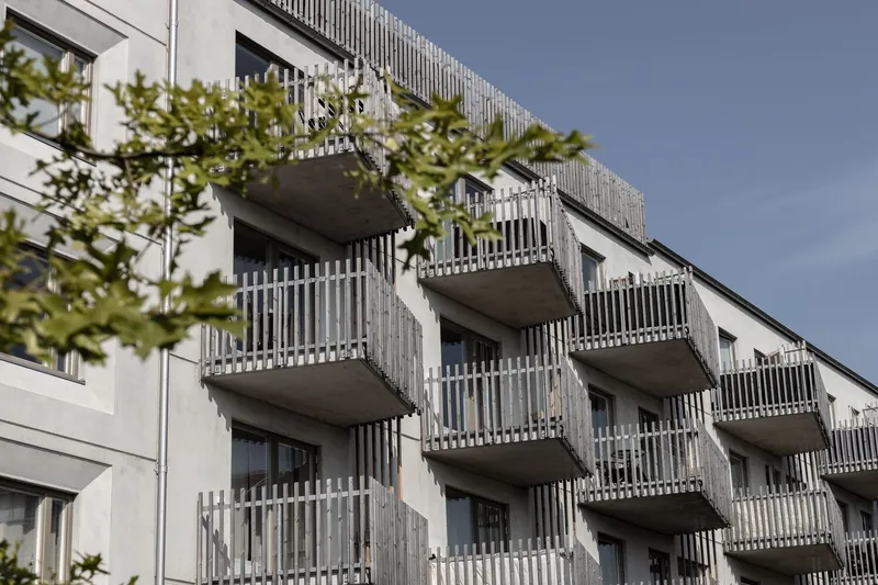 Facade with stacked concrete balconies and vertical white screening elements viewed through green foliage
