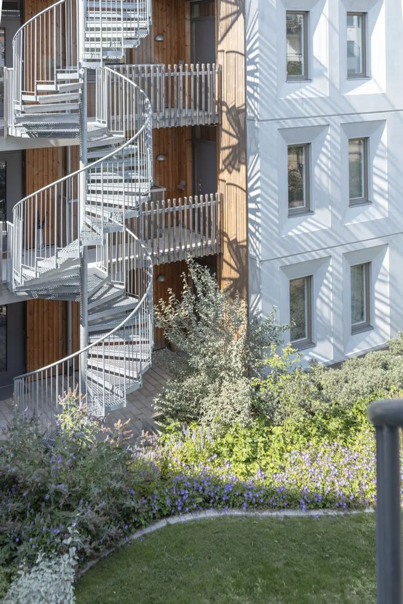 Metal spiral staircase connecting timber-clad balconies above a planted courtyard with flowering vegetation