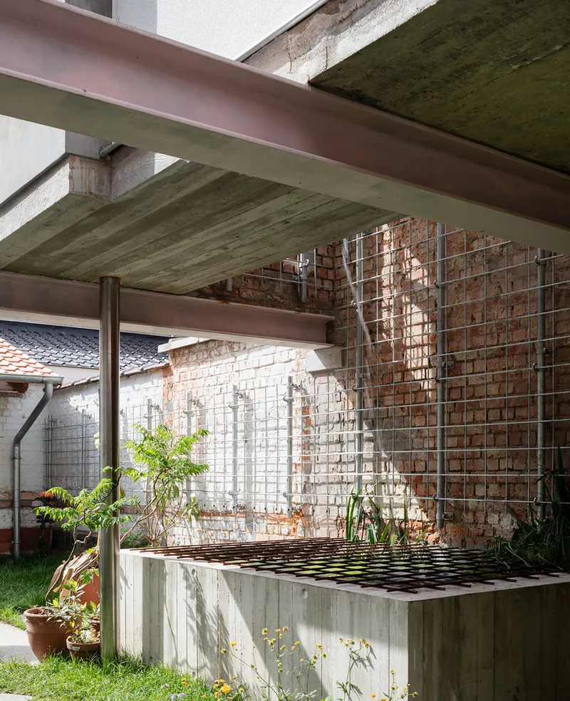 Covered courtyard with exposed concrete beams, wire mesh screen, and planted beds along a brick wall