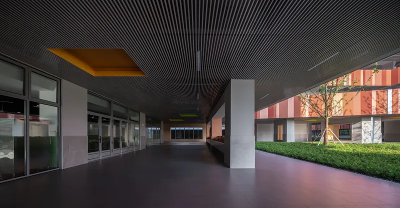 Covered corridor with dark slatted ceiling and square columns leading toward illuminated courtyard facades at dusk