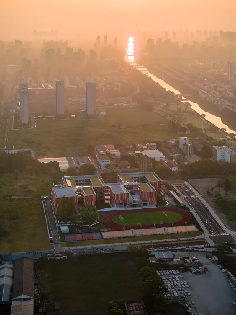 Aerial view of the school campus with vertical striped cladding and green rooftop sports field at golden hour