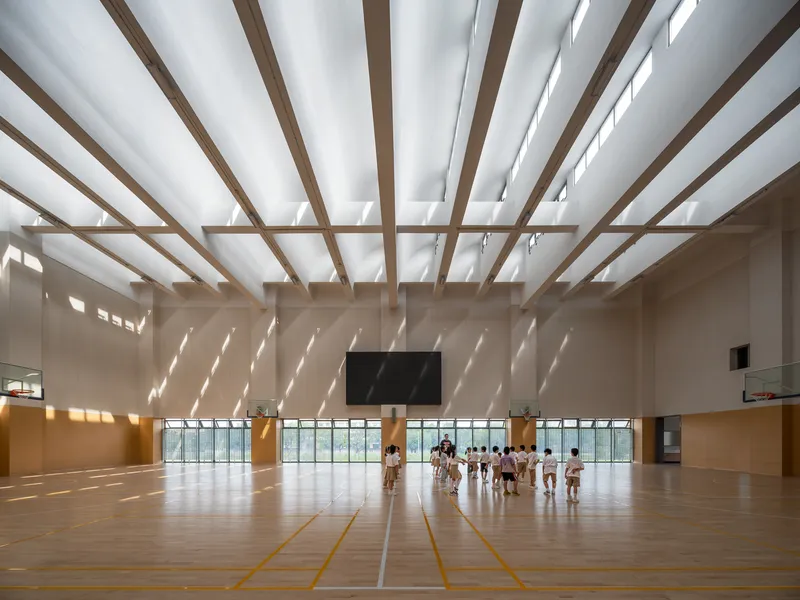 Interior sports hall with timber louvered ceiling casting diagonal shadows across students practicing on wooden floor