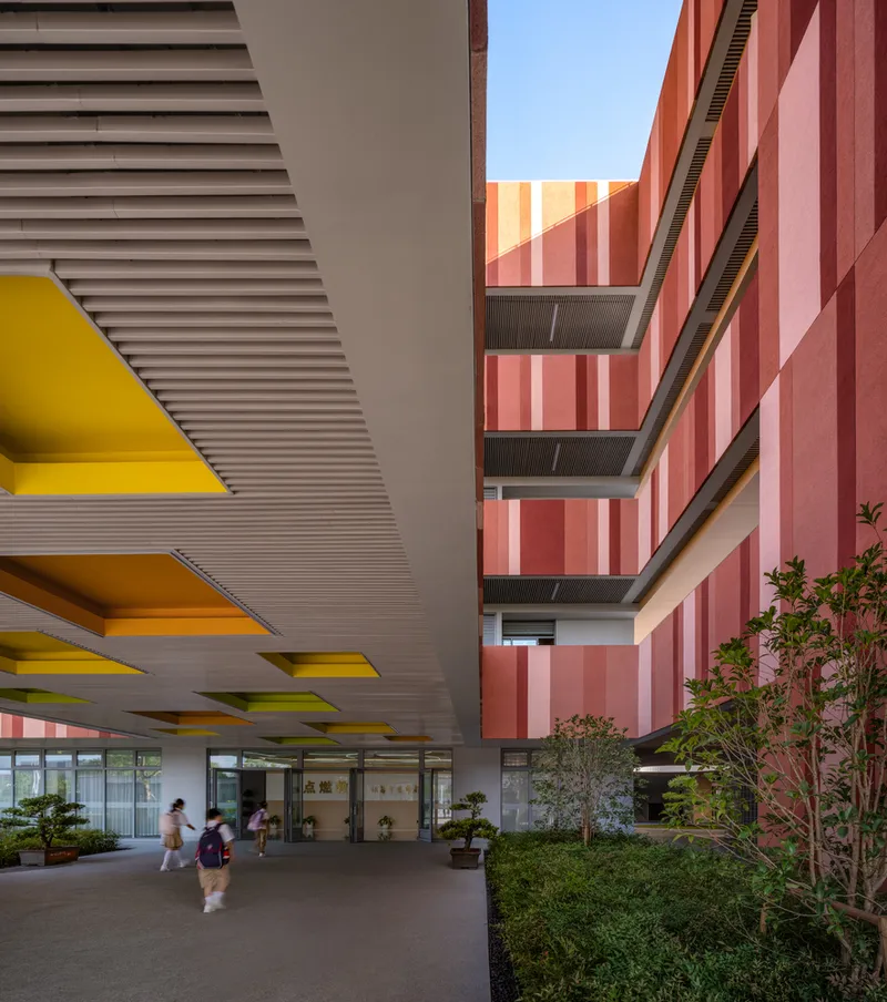 Covered entry passage with yellow triangular ceiling coffers between striped facade volumes and planted courtyard