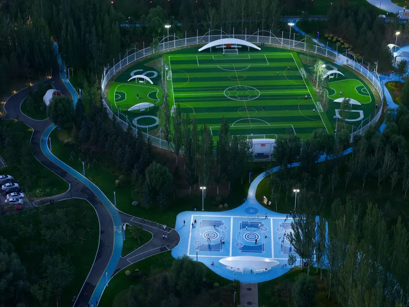 Overhead view at dusk showing illuminated basketball court and striped football pitch connected by flowing pathways