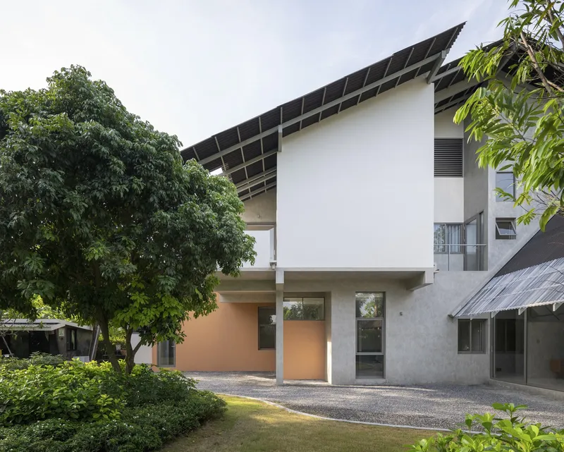 Two-story facade with asymmetrical window openings and sloped roof visible between mature trees