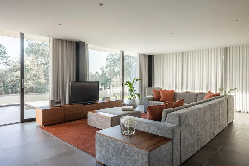 Living room with grey sectional sofa and floor-to-ceiling curtains framing glazed walls overlooking trees