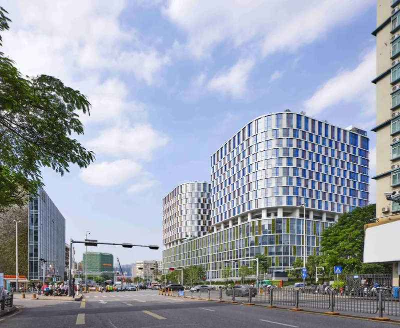 Street view of two curved glass towers with chequered facades flanked by trees and traffic barriers