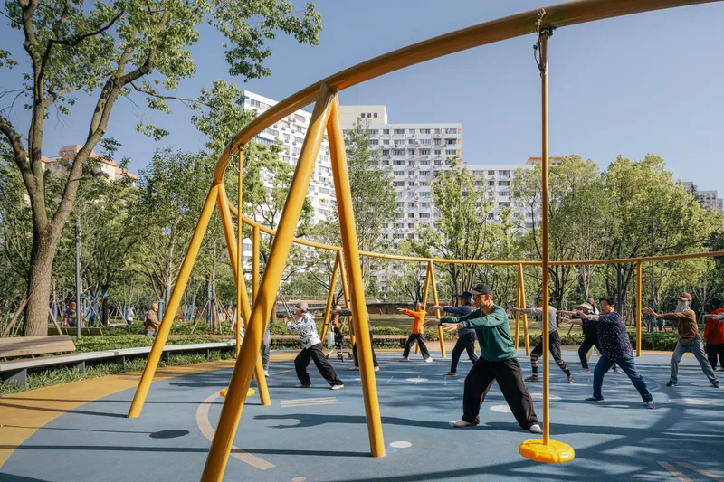 Yellow steel exercise frame sheltering group activity beneath apartment towers and tree canopy
