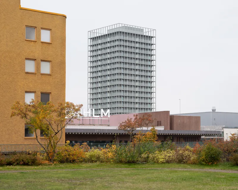 Tall residential tower with repetitive balcony facade rising above low-rise buildings and autumn foliage in an overcast day