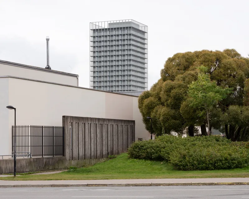 Low plaster building with vertical timber cladding beside a hedge and cluster of mature trees under grey skies