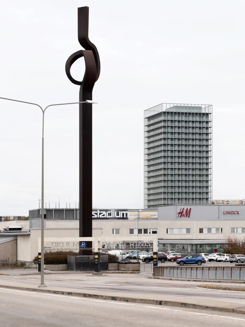 Black sculptural monument beside a roadway with a repetitive facade tower in the background