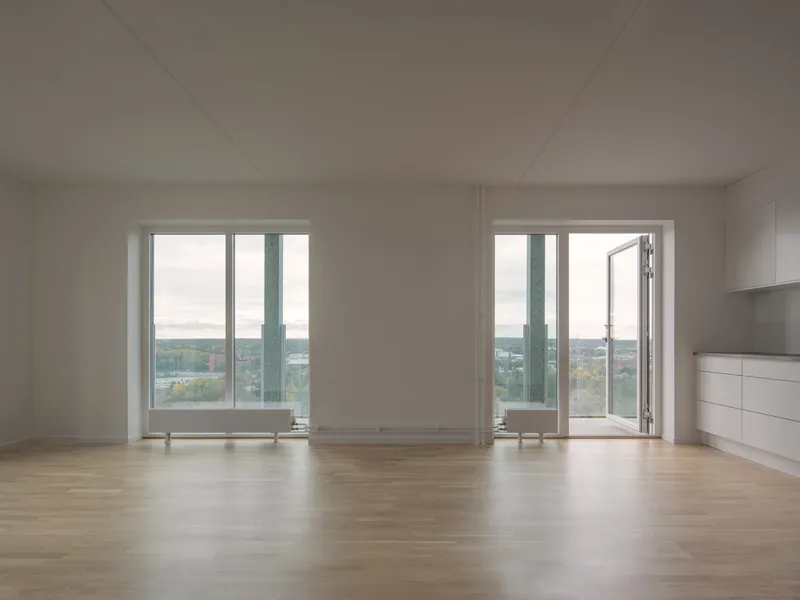 Empty interior room with light timber flooring and two floor-to-ceiling windows overlooking distant rooftops