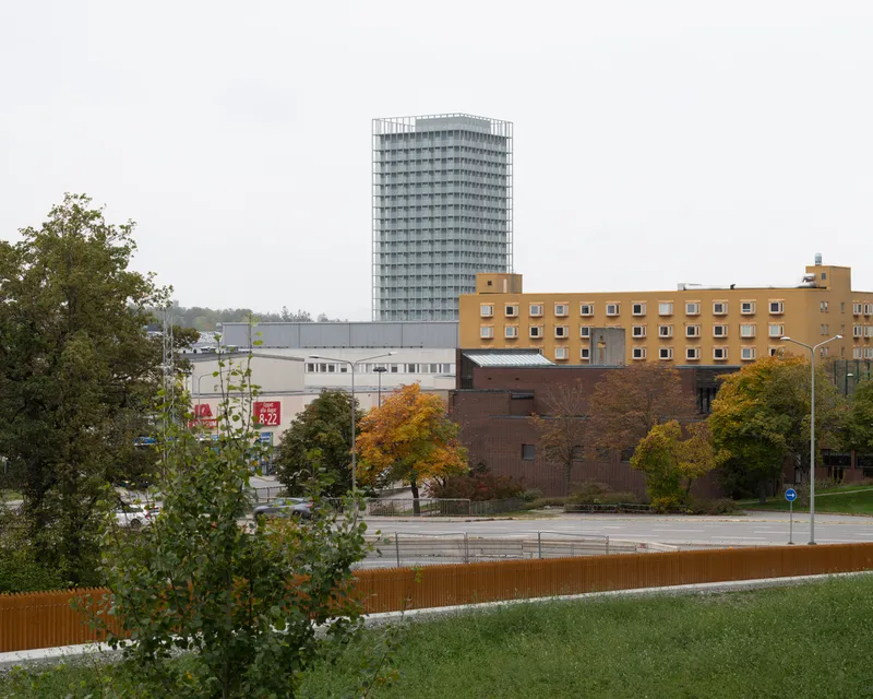 High-rise tower with gridded facade rising above brick structures and autumn trees